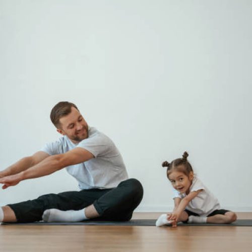 Teaching how to stretch the legs. Father with little daughter are doing yoga at home.